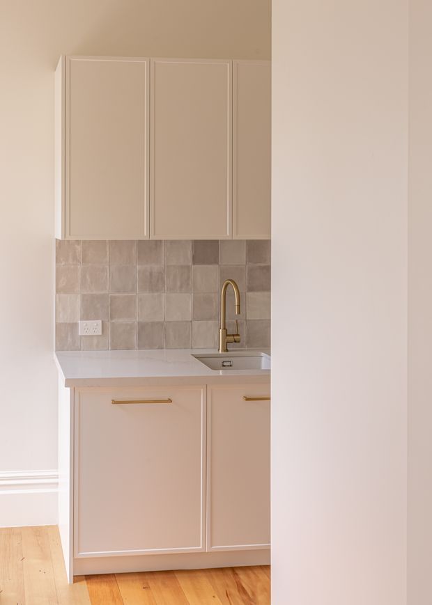 A compact, softly styled kitchenette viewed from an adjacent room or hallway. The space features pale blush-beige shaker-style upper and lower cabinetry with slim gold bar pulls, a white engineered-looking benchtop, a small stainless-steel sink with a brass gooseneck tap, and a square taupe-grey tiled splashback. Warm timber flooring and white skirting boards complete the understated fit-out.