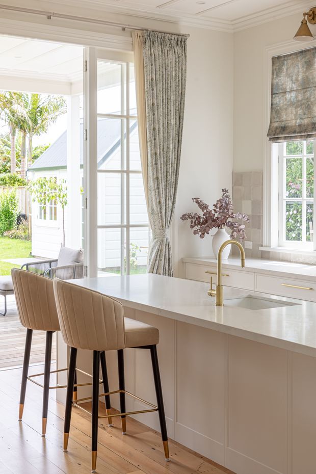 Bright, refined kitchen interior with a large pale stone island, integrated sink and brushed brass gooseneck tap, cream shaker-style cabinetry, timber flooring, and two upholstered bar stools. Tall French doors open to a small outdoor deck and garden, while a window with a patterned Roman blind and full-height curtains brings in abundant natural light. The room has decorative ceiling moulding and a soft neutral palette with brass accents.