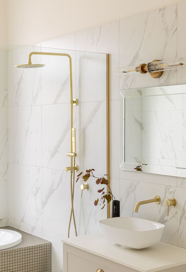 Bright, refined bathroom featuring white marble-look wall tiles, a frameless mirrored cabinet, a white vessel basin on a pale vanity, and coordinated brushed brass shower, tapware, and lighting. A tiled bath or ledge is visible at lower left, with a small decorative plant adding warmth.