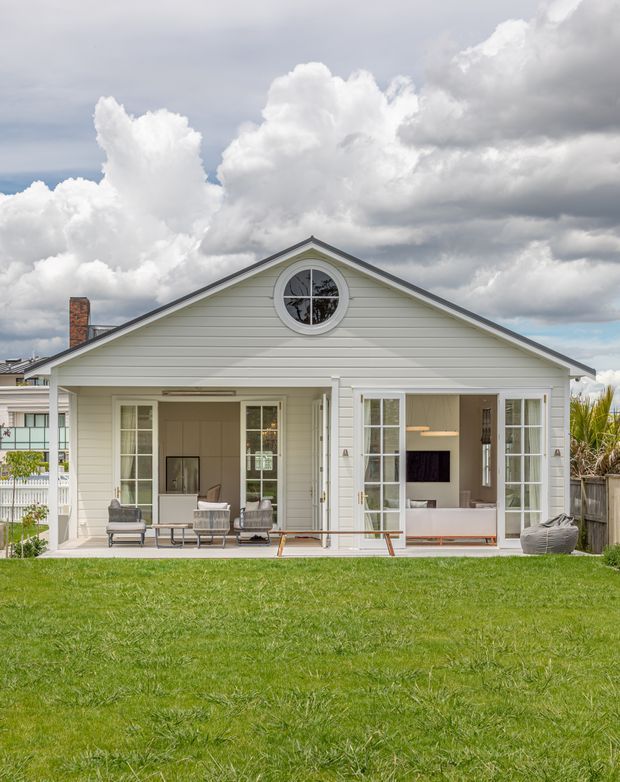 Front exterior view of a single-storey weatherboard home with a symmetrical gabled facade, circular attic window, covered outdoor living area, and large glazed openings. The building is painted pale off-white, with a brick chimney and a broad lawn in the foreground under a dramatic cloudy sky.