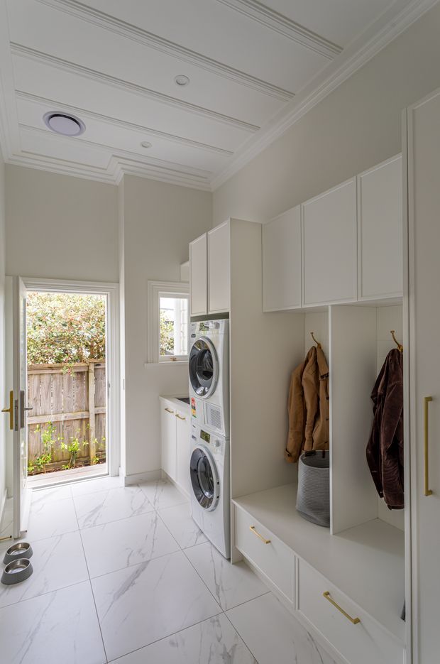 Bright, narrow laundry and utility room with white built-in cabinetry, stacked front-loading washer and dryer, marble-look floor tiles, a decorative coffered ceiling, brass hardware, and an integrated bench with open hanging compartments. A glazed exterior door and window provide natural light and views to a fenced garden area.