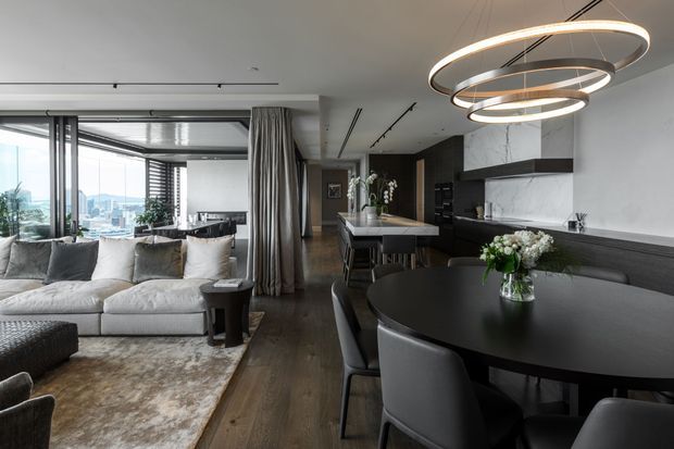 Open-plan contemporary apartment interior combining a living room, dining area, and kitchen. The space features floor-to-ceiling glazing with city views, a pale sectional sofa, dark round dining table, dark kitchen cabinetry, marble-look wall surfaces, timber flooring, layered curtains, and sculptural circular pendant lighting.