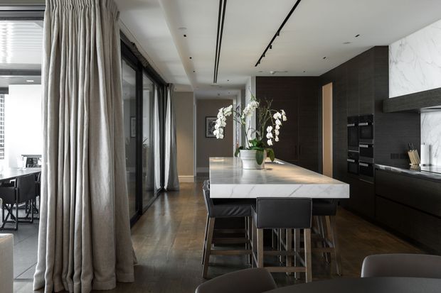Contemporary open-plan kitchen and dining area with a long marble island, dark timber cabinetry, integrated appliances, black track lighting, and floor-to-ceiling glazing screened by full-height neutral curtains. A dining table is visible at the left, while orchids and stools occupy the island.