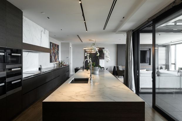 A spacious contemporary kitchen centered on a large marble-effect island with an integrated sink and dark wood base. The kitchen features extensive dark cabinetry, built-in appliances, a marble-look splashback, linear recessed and track lighting, and a dining area beyond. Full-height black-framed glazed doors open toward a bright living space, creating a refined open-plan interior.