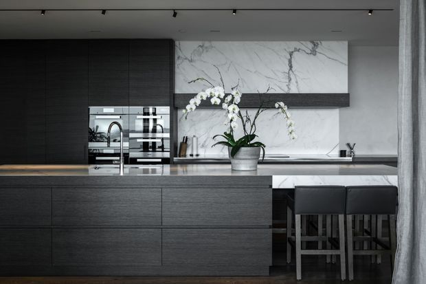 A sophisticated contemporary kitchen featuring dark timber cabinetry, a large central island with integrated sink and tapware, built-in stainless-steel appliances, and a dramatic white marble-look splashback and wall panel. Black bar stools sit at the island, with understated track lighting and a potted orchid adding a refined touch.