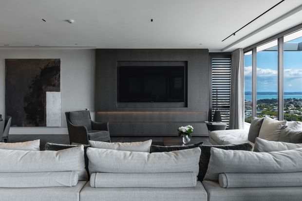 Contemporary living room with a large light-grey sectional sofa facing a dark built-in media wall and television. Floor-to-ceiling corner glazing provides expansive coastal and urban views, while neutral upholstery, dark timber-toned joinery, and minimal furnishings create a refined, modern atmosphere.