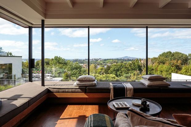 A contemporary living room with expansive floor-to-ceiling glazing overlooking a leafy suburban landscape and distant hills. A built-in timber-framed window seat or sectional bench runs along the glazing, with cushions and a small round coffee table in the foreground. The ceiling has exposed timber beams, and the room receives abundant natural light.