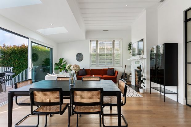 Bright, open-plan dining and living area with a black-framed rectangular dining table surrounded by woven cane-back chairs. The room features warm timber flooring, white walls, a red-orange sofa, a white fireplace mantel, indoor plants, large glazed doors opening to a deck, and a white slatted ceiling with skylights. Black-framed glazing and a tall black display cabinet add contrast.