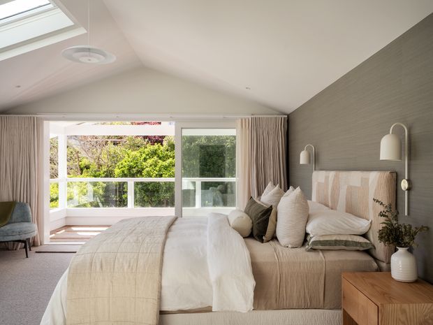 Bright, calm contemporary bedroom with a pitched vaulted ceiling and skylight. The room features a large upholstered bed with layered neutral bedding and textured pillows, a grey textured feature wall, fluted wall-mounted bedside lamps, timber bedside cabinetry, soft carpet, and full-height glazed doors opening to a balcony or leafy outdoor view. Neutral linen, cream, beige, and grey tones create a refined, relaxed interior.