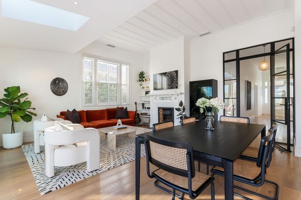 Bright open-plan living and dining area with white walls, light timber flooring, a black dining table with woven-seat chairs, and a white lounge setting on a patterned rug. The room features a traditional fireplace with mantel, large windows with blinds, a skylight, white painted ceiling panelling, indoor plants, and black-framed glazed doors opening to a hallway.