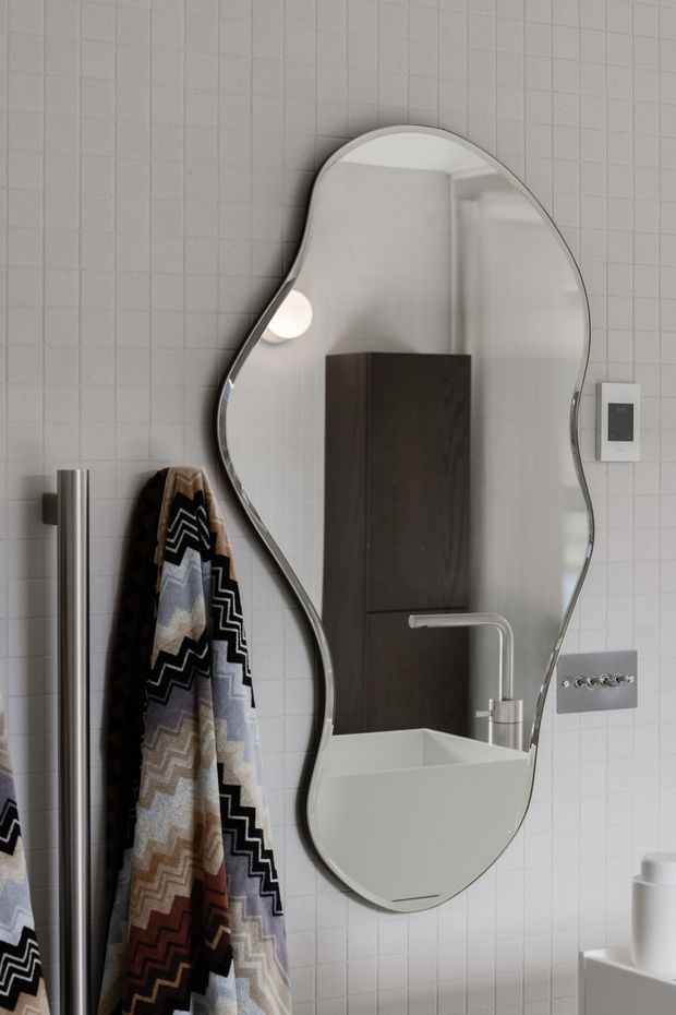 A close-up of a contemporary bathroom featuring a large organically shaped frameless mirror with a polished edge, mounted over a white tiled wall. The mirror reflects a white basin with a slim chrome tap and dark timber vanity cabinetry. A patterned towel hangs beside a tall cylindrical chrome towel rail, with minimalist wall controls and fixtures visible to the right.