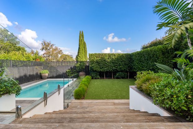 A landscaped residential backyard viewed from a broad timber staircase and deck. The scene includes a compact swimming pool with glass safety fencing on the left, a manicured lawn bordered by dense hedges, raised white-rendered garden beds filled with tropical and ornamental planting, and a tall narrow cypress tree. Bright blue sky and lush greenery create a polished outdoor entertaining and garden setting.