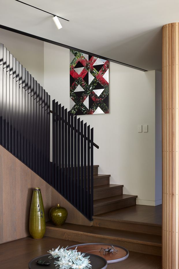 Interior view of a contemporary stairway and landing with dark timber treads and flooring, a black vertical metal balustrade, warm wood wall panelling, and a curved timber-clad column. Neutral white walls, linear track lighting, and a bold geometric botanical artwork complete the refined modern composition. Decorative vases and trays sit at the base of the stairs.