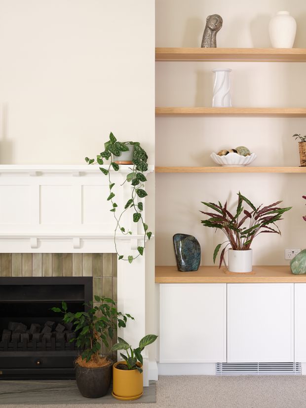 Bright, calm interior detail showing a traditional-style fireplace with white panelled mantel and a built-in shelving and cabinetry unit. Open timber shelves display ceramics, stones and indoor plants, while white lower cabinets provide concealed storage. The fireplace surround uses vertically arranged muted green-grey tiles, and the floor is carpeted in a light neutral tone.
