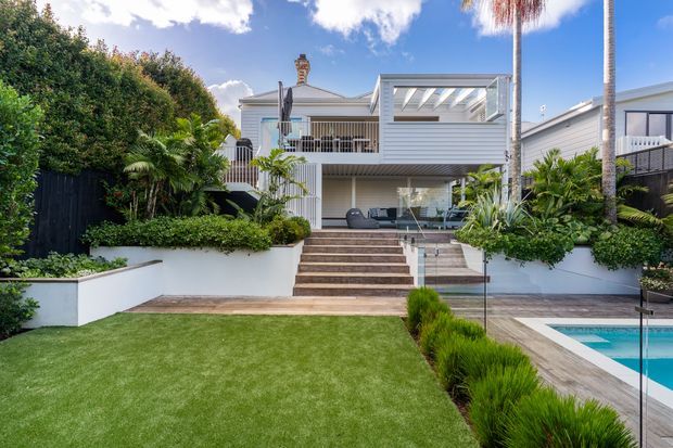 A bright rear exterior view of a contemporary coastal-style home with white horizontal cladding, a raised timber deck and balcony, broad outdoor steps, landscaped garden beds, and a swimming pool in the foreground. Glass balustrades and a covered pergola create a seamless connection between the house and pool terrace. Tall palms and lush planting contribute to a resort-like setting.