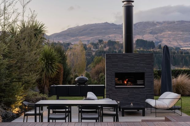 Outdoor dining and lounge area on a timber deck, overlooking a mountainous landscape. The space features a long dining table with black chairs, a built-in dark masonry fireplace with a tall chimney, a compact outdoor grill, and a sculptural lounge chair. Dense landscaped planting frames the terrace.