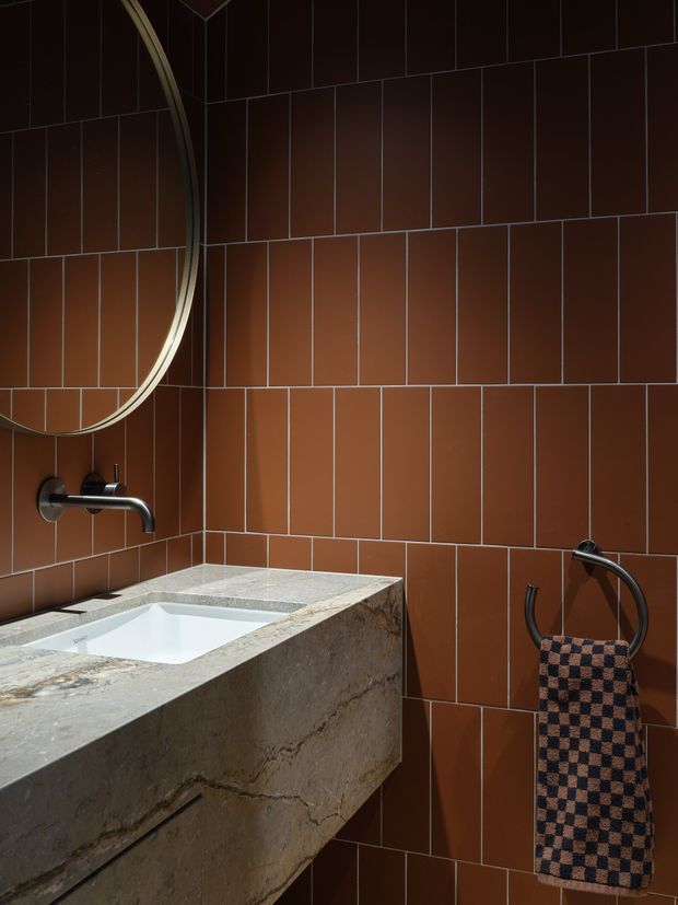 A moody bathroom vanity area clad in rich terracotta-brown rectangular tiles with contrasting grout. The image shows a large round mirror, a wall-mounted brushed metal tap, and a substantial stone vanity with an integrated white basin. A dark metal towel ring with a patterned towel is mounted on the tiled wall.