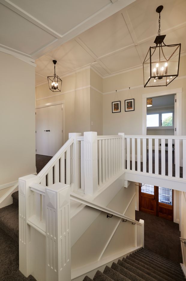 A bright, traditional multi-level stairway and landing with white painted timber balustrades, panelled walls, decorative ceiling mouldings, and geometric black pendant lights. The carpeted staircase descends toward a timber front door, while an upper landing and adjoining rooms are visible.