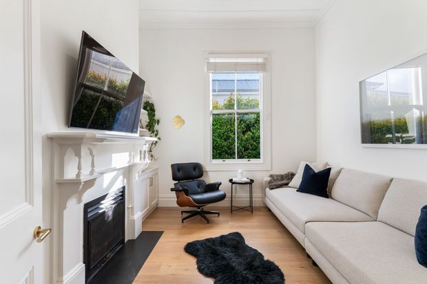 Bright, compact living room with white walls, decorative ceiling cornice, timber flooring, a traditional white mantel fireplace, wall-mounted television, contemporary sectional sofa, black leather lounge chair, and a central dark sheepskin rug. A window brings in natural light and reflects greenery outside. The room blends traditional architectural detailing with contemporary furnishings.