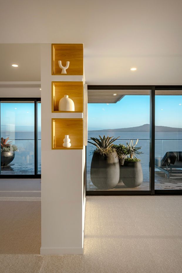 Bright contemporary interior overlooking a body of water and distant hills through expansive black-framed glazing. A central white structural or partition column contains three recessed timber display niches with sculptural ceramics. The room has warm neutral carpet, a smooth white ceiling with recessed downlights, and access to an outdoor deck visible through the glazing. Large planters and outdoor seating appear beyond the windows.