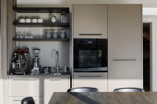 A compact, contemporary kitchen viewed from the dining area. The space features flat-panel light neutral cabinetry, integrated black appliances, open dark timber shelving with coffee and tea accessories, a stone-look benchtop, and a stainless steel sink tap. A dining table and chairs occupy the foreground, with a doorway and additional cabinetry visible at right.