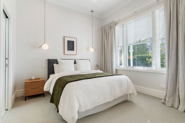 Bright, refined bedroom with a neatly made upholstered bed, white bedding, olive-green throw, timber bedside table, pendant lights, framed artwork, and a large window fitted with white blinds and full-length curtains. The room has pale walls, carpeted flooring, simple decorative moulding, and an airy contemporary feel.