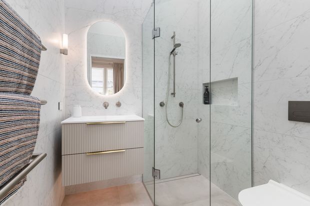 Bright contemporary bathroom with full-height pale marble-look wall cladding, a frameless glass walk-in shower, floating fluted vanity with brass hardware, wall-mounted basin, rounded backlit mirror, and concealed toilet cistern. A heated towel rail and recessed shower niche are visible.