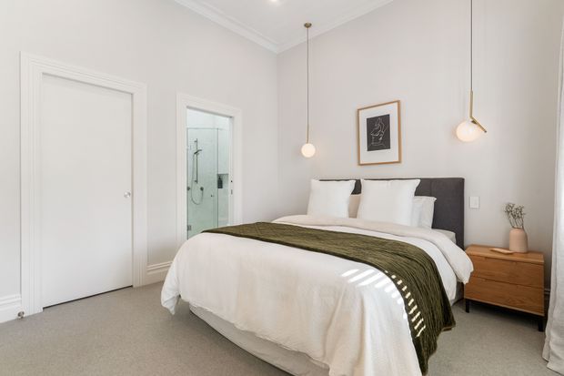 A bright, refined bedroom with a neatly made bed, white bedding, dark upholstered headboard, and an olive-green throw. The room has pale grey walls, white decorative cornices and door trim, soft carpeting, minimalist pendant lights, framed artwork, and a timber bedside table. A glazed shower area is visible through an open doorway, suggesting an adjacent bathroom.