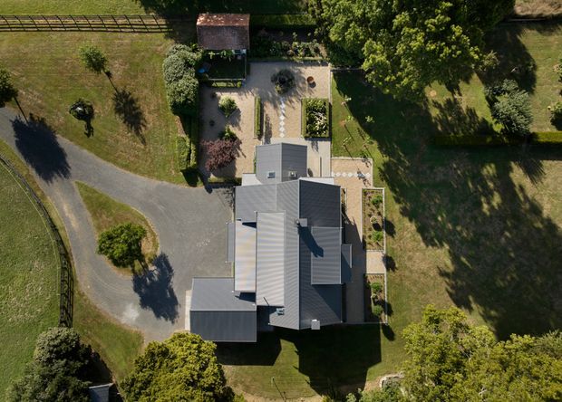 Aerial top-down view of a large detached residential property set within expansive landscaped grounds. The house has a complex dark grey pitched roof with multiple connected volumes, surrounded by lawns, mature trees, paved garden terraces, planted beds, and a curved driveway leading to the property.
