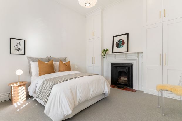 Bright, refined bedroom with a neatly made upholstered bed, white bedding, grey and ochre accent cushions, built-in white panelled wardrobes, and a decorative traditional fireplace with ornate mantel. The room includes framed artwork, a small bedside table with lamp, a sculptural ceiling light, and a pale yellow accent chair. Overall styling is calm and contemporary with classic architectural detailing.