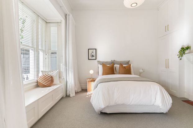 Bright, softly styled bedroom with a centered upholstered bed dressed in white bedding and layered neutral and tan cushions. The room features a large bay-style window with white blinds and full-height curtains, a built-in window seat with drawers, extensive white built-in wardrobes, pale carpet, bedside tables, and restrained wall art. The overall look is calm, airy, and contemporary with traditional detailing such as wall and ceiling mouldings.