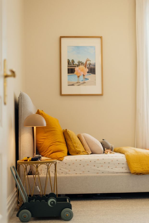 A warmly styled children's bedroom corner featuring a low upholstered bed or daybed, layered mustard yellow and neutral cushions, a matching yellow throw, a small bedside table with a dome lamp, framed poolside artwork, and a dark wheeled children's toy in the foreground. The room has soft cream walls, light carpet, and a calm, playful atmosphere.