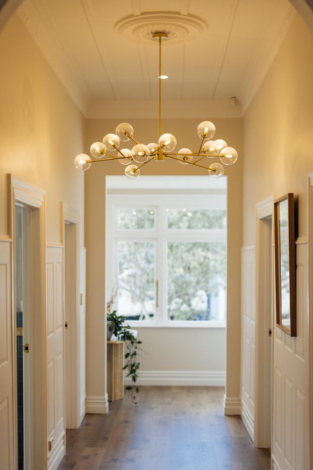 A warm, elegant residential hallway with painted cream walls, white decorative wainscoting and moulded door trims, a timber floor, and a large brass chandelier with spherical glass bulbs. The corridor leads to a bright windowed area at the rear, with mirrors and doorways along both sides. An ornate ceiling medallion and panelled ceiling add traditional architectural detail.
