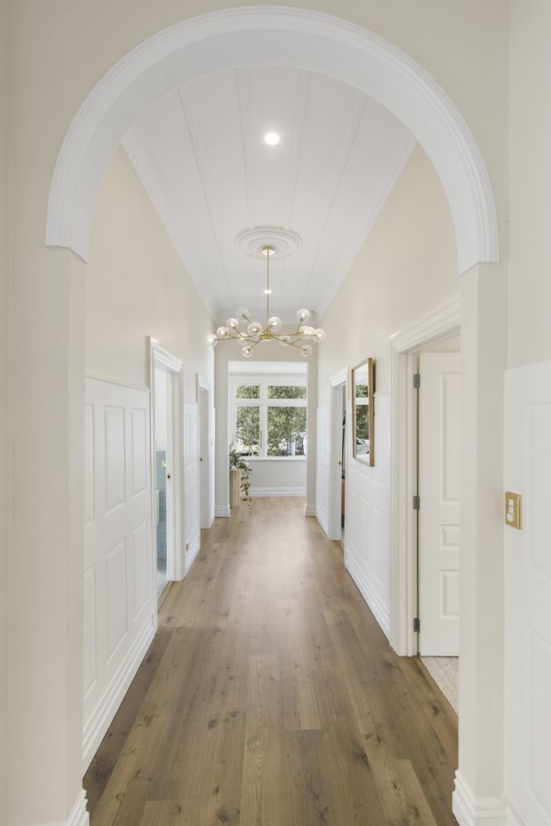 Bright, elegant residential hallway viewed through a large rounded archway. The corridor has white paneled doors and wall wainscoting, ornate white ceiling detailing, a contemporary branching chandelier, pale timber flooring, and a window at the far end.