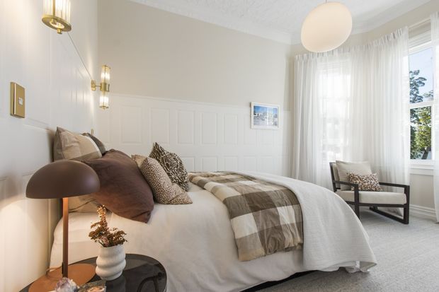 Bright, refined bedroom with a neatly made bed layered in white bedding, brown and patterned cushions, and a taupe-and-cream checked throw. The room features white decorative wall panelling, sheer floor-to-ceiling curtains, a large window, wall sconces, a sculptural pendant light, a dark bedside table with lamp, and a modern upholstered armchair. The overall look is warm, elegant, and softly contemporary.