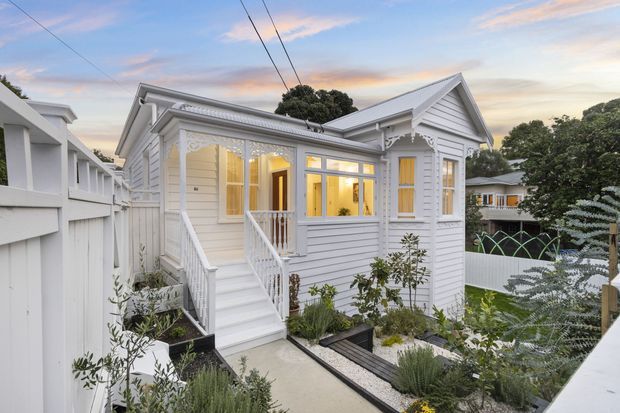 Exterior view of a compact raised white weatherboard cottage with a gabled roof, ornate decorative fretwork, sash-style windows, and a small covered entry porch reached by stairs. A landscaped front garden and paved path surround the house, with neighbouring homes visible beyond.