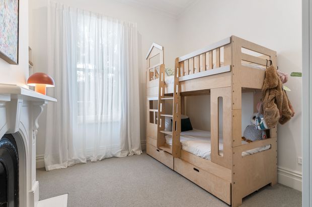 Bright, compact children's bedroom with a built-in or fitted timber bunk bed featuring integrated ladder, guardrails, storage drawers and shelving. The room has pale walls, light carpet, sheer floor-to-ceiling curtains, traditional white decorative fireplace detailing, and a small orange table lamp. Soft toys and children's belongings are visible around the bunk, creating a calm, practical and playful atmosphere.