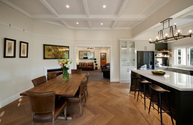 A refined open-plan dining and kitchen interior. The foreground features a large timber dining table with upholstered leather dining chairs, while the kitchen has a substantial light stone island with dark cabinetry, integrated seating, open shelving, and white-framed windows. A coffered ceiling with recessed lighting spans the room, and a decorative linear pendant hangs above the island. Herringbone timber flooring, framed artwork, built-in cabinetry, and a glimpse of a living room beyond complete the warm, traditional-contemporary space.