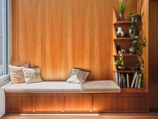 Built-in timber window seat and cabinetry forming a warm reading nook, with a cushioned bench, pillows, open display shelving, books, ceramics, and trailing plants. Vertical wood panelling and joinery create a richly detailed, cozy interior feature.