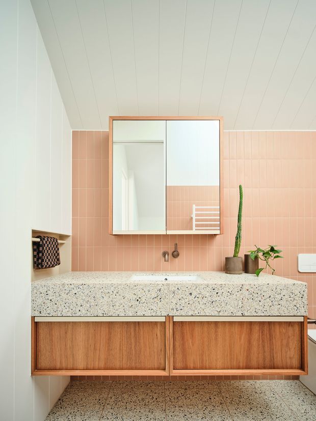 A bright, compact bathroom vanity with a wall-mounted double cabinet in warm timber veneer, a terrazzo countertop and matching terrazzo floor tiles. The backsplash is clad in soft pink rectangular tiles, with a framed mirrored medicine cabinet above the integrated basin. White vertical wall panelling and a sloped panelled ceiling create a clean, contemporary feel, while small potted plants add a natural accent.
