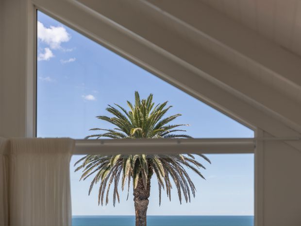 Interior view through large angled floor-to-ceiling glazing, with a palm tree and blue ocean horizon visible outside. The room has a clean, light-toned architectural envelope with white window frames, a horizontal structural or glazing bar, and a pale curtain at the left. The image feels bright, airy, and coastal.