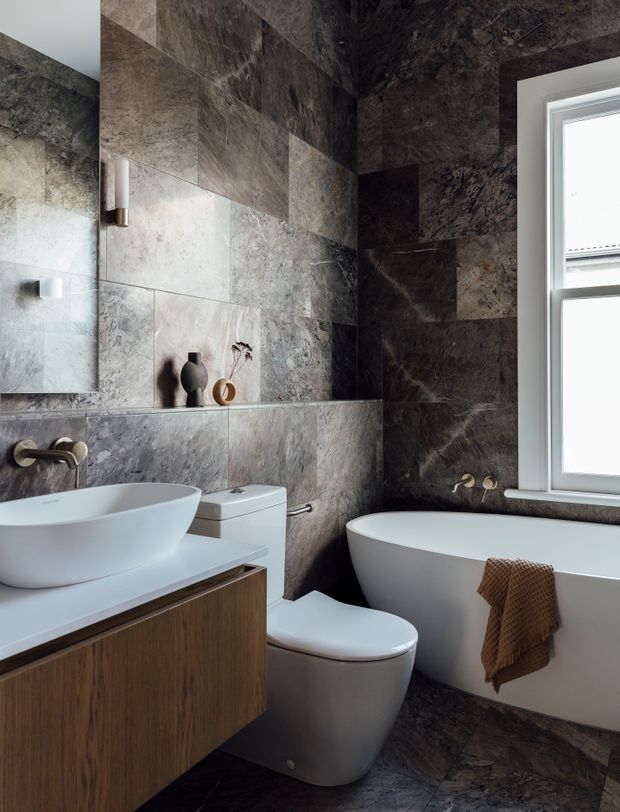 A compact, refined bathroom clad in large-format dark veined stone-look tiles across the walls and floor. The room features a freestanding white bathtub beneath a window, a wall-hung white toilet, and a white vessel basin set on a warm timber floating vanity. Minimal ceramic and glass decor sits on a recessed ledge, with brushed metal tapware and soft natural light.