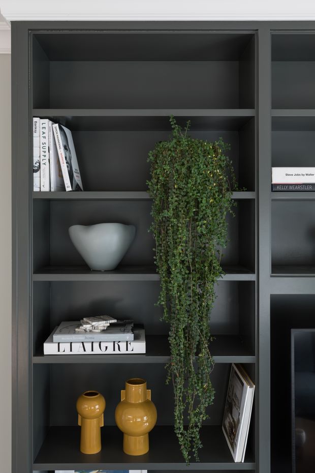 Close-up interior view of dark charcoal built-in shelving with open compartments, styled with books, a pale sculptural vase, trailing greenery, mustard-yellow ceramic vessels, and framed artwork. The shelving is set beneath crisp white ceiling cornices, creating a refined, moody display feature.