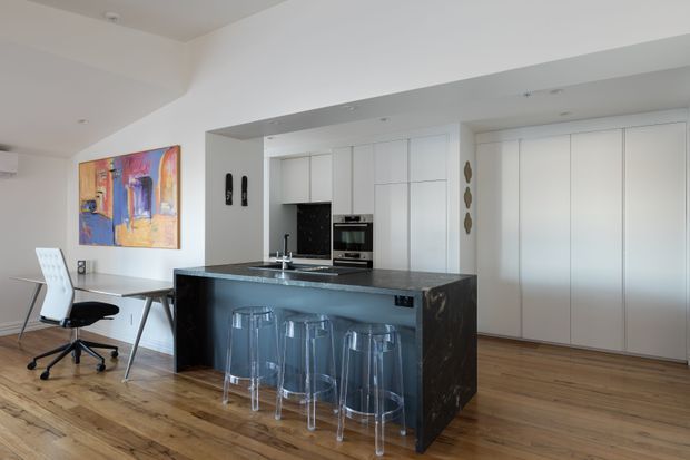Open-plan contemporary kitchen and dining/work area with a large dark stone island, white handleless cabinetry, integrated appliances, clear bar stools, timber flooring, and a compact desk beneath colourful wall art. The space is bright and uncluttered with a restrained neutral palette and a bold artwork accent.