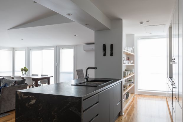 Bright open-plan kitchen viewed from the island, with dark stone benchtops, grey built-in cabinetry, an integrated sink and tap, open timber shelving, and light timber flooring. The space opens to a dining and living area with extensive windows and white roller blinds.