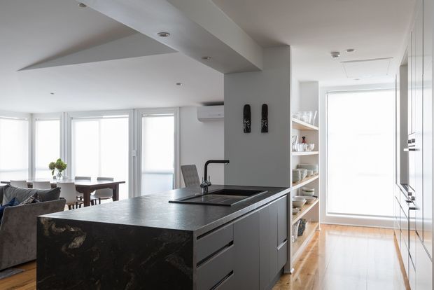 Bright open-plan contemporary interior viewed from the kitchen, featuring a large dark stone island with integrated sink and tap, streamlined grey cabinetry, open shelving, timber flooring, and a dining and living area beyond. Floor-to-ceiling glazing and white finishes create an airy, light-filled space.