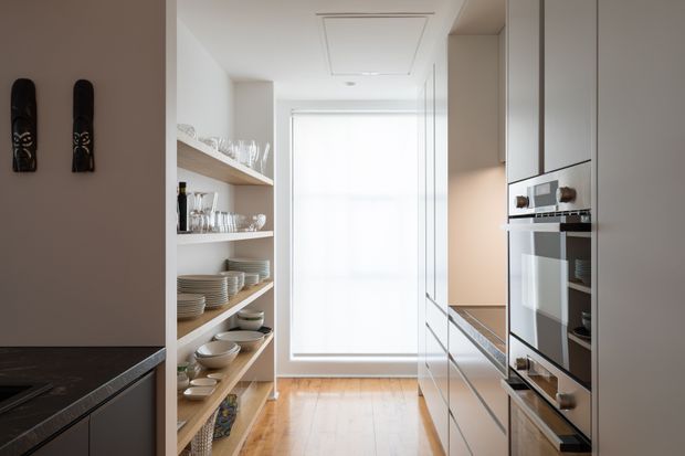 Bright, narrow contemporary kitchen with a walk-in pantry corridor. Open timber-toned shelving displays dishes and glassware on the left, while sleek pale cabinetry with integrated appliances and a dark benchtop lines the right. A large translucent window at the end brings in soft natural light, and light timber flooring runs through the space.