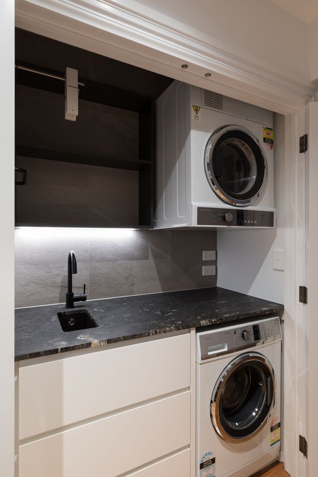A compact, contemporary laundry room with stacked front-loading washer and dryer, dark cabinetry, a black stone-look benchtop, integrated black sink and tap, tiled splashback, and under-cabinet lighting. White cabinetry and decorative white door trim create contrast with the dark finishes.