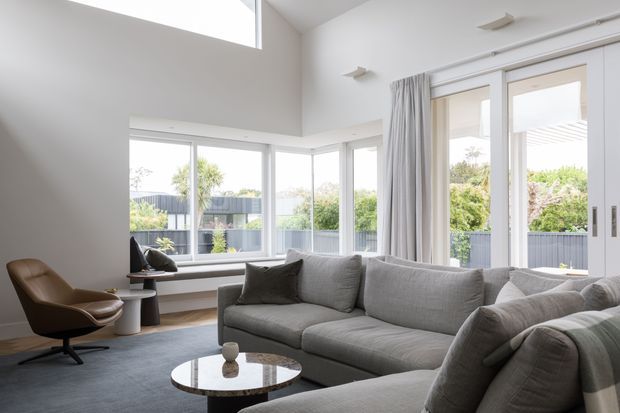 Bright contemporary living room with a large grey sectional sofa, sculptural brown lounge chair, round coffee table, and expansive corner glazing. White walls, high ceilings, sheer curtains, and glazed doors create an airy connection to the garden and outdoor terrace.