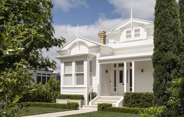 A bright exterior view of a refined white weatherboard house with Victorian-era detailing, including decorative gables, classical trim, louvred shutters, a projecting bay window, front steps, and a prominent chimney. Formal hedging, clipped shrubs, mature trees, and a paved path frame the facade.